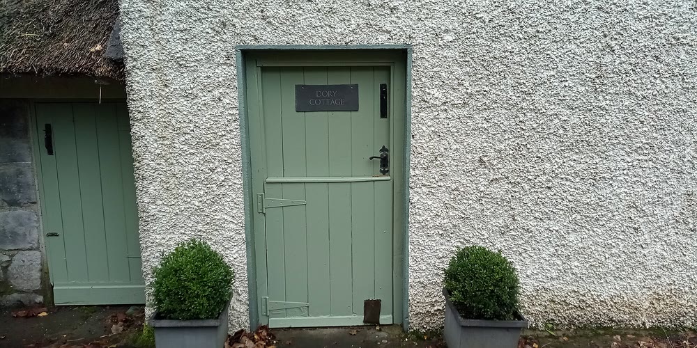 A pale green wooden half-door with a slate name plate and black latch set in a white pebble-dashed cottage wall, with clipped box plants in grey planters on either side and the edge of a thatched roof at the left