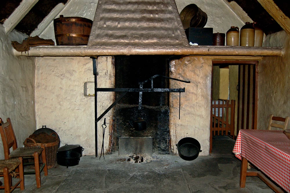 An open hearth under a wide canopy chimney in an old farmhouse kitchen, with an iron crane, black pots on the flagstone floor, straw-seated chairs at the left and a table with a red check cloth at the right