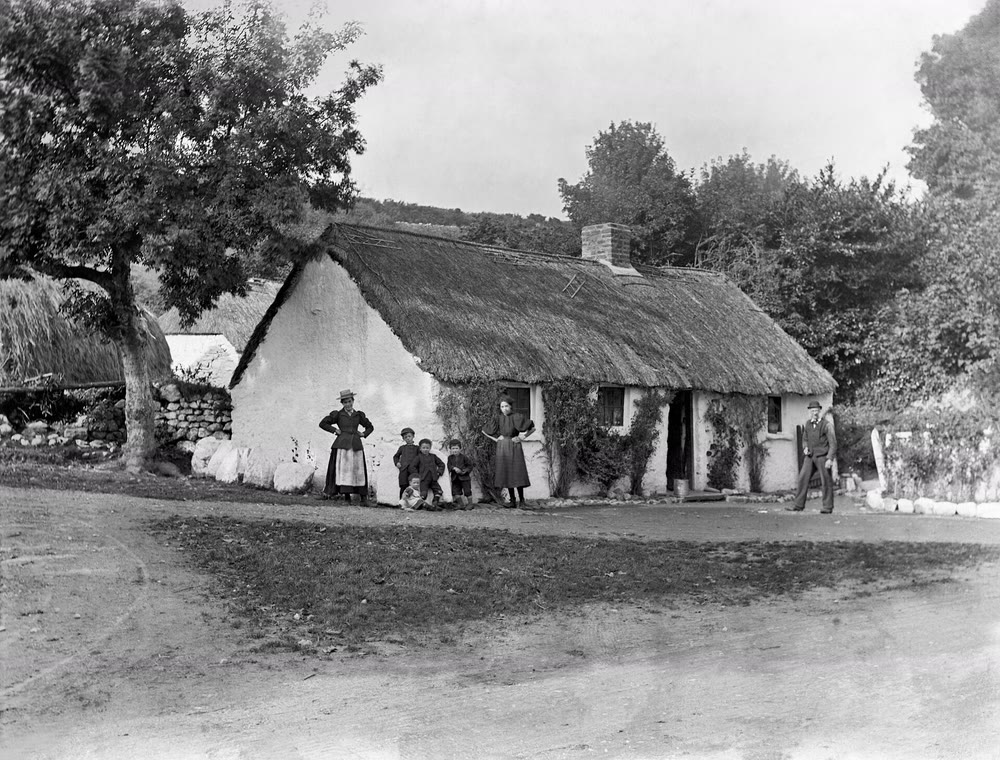 Black-and-white photograph from about 1895 of a long whitewashed thatched cottage with a chimney and open door, two women and several children standing in front, a man at the right and a large tree at the left