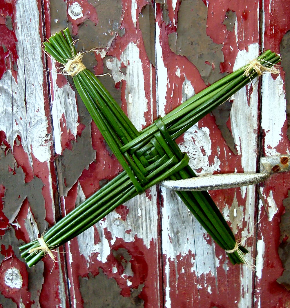 A green rush St Brigid's cross with a woven square centre and four tied arms, fixed to an old wooden door with flaking red and grey paint and a metal latch