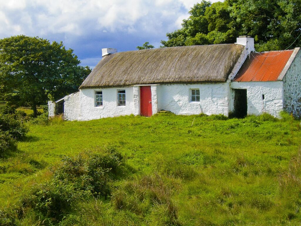 A long whitewashed thatched cottage with a red front door and small windows in a field of rough green grass in Co. Donegal, with a red corrugated-roofed outhouse at the right and trees under a cloudy blue sky