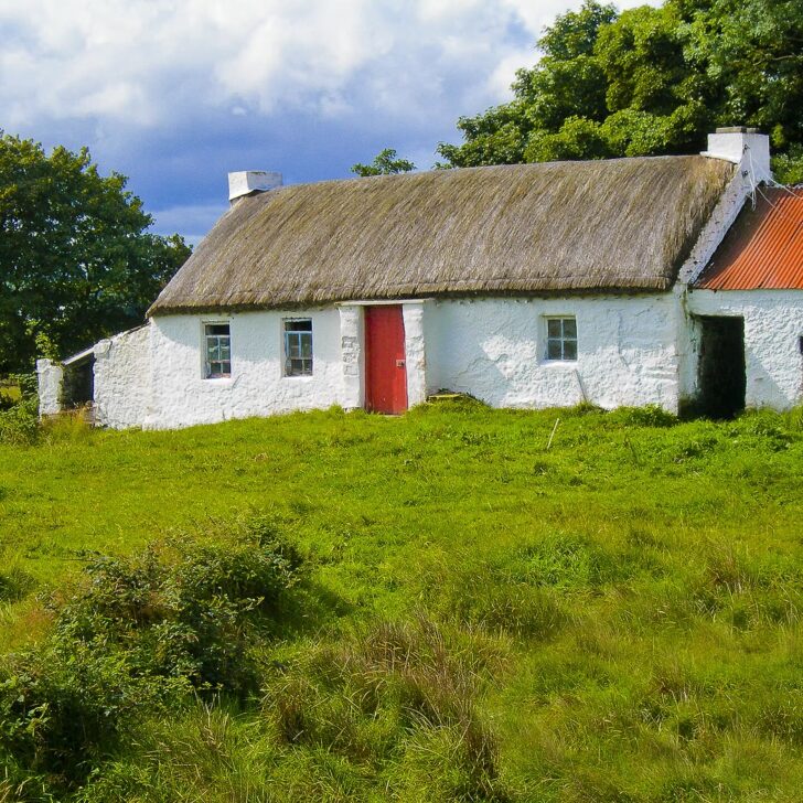 A long whitewashed thatched cottage with a red front door and small windows in a field of rough green grass in Co. Donegal, with a red corrugated-roofed outhouse at the right and trees under a cloudy blue sky