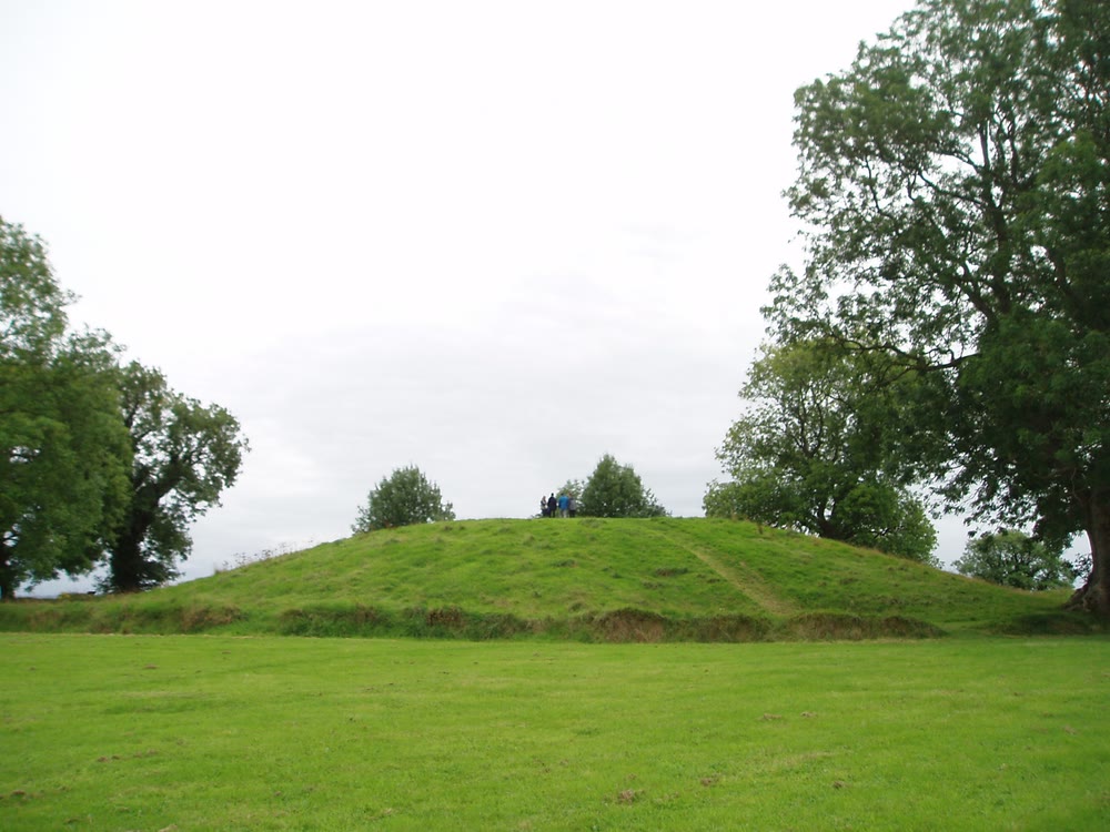 A broad grassy mound with a worn path up its side, flanked by tall trees under a pale sky, with a few tiny distant figures standing on the top