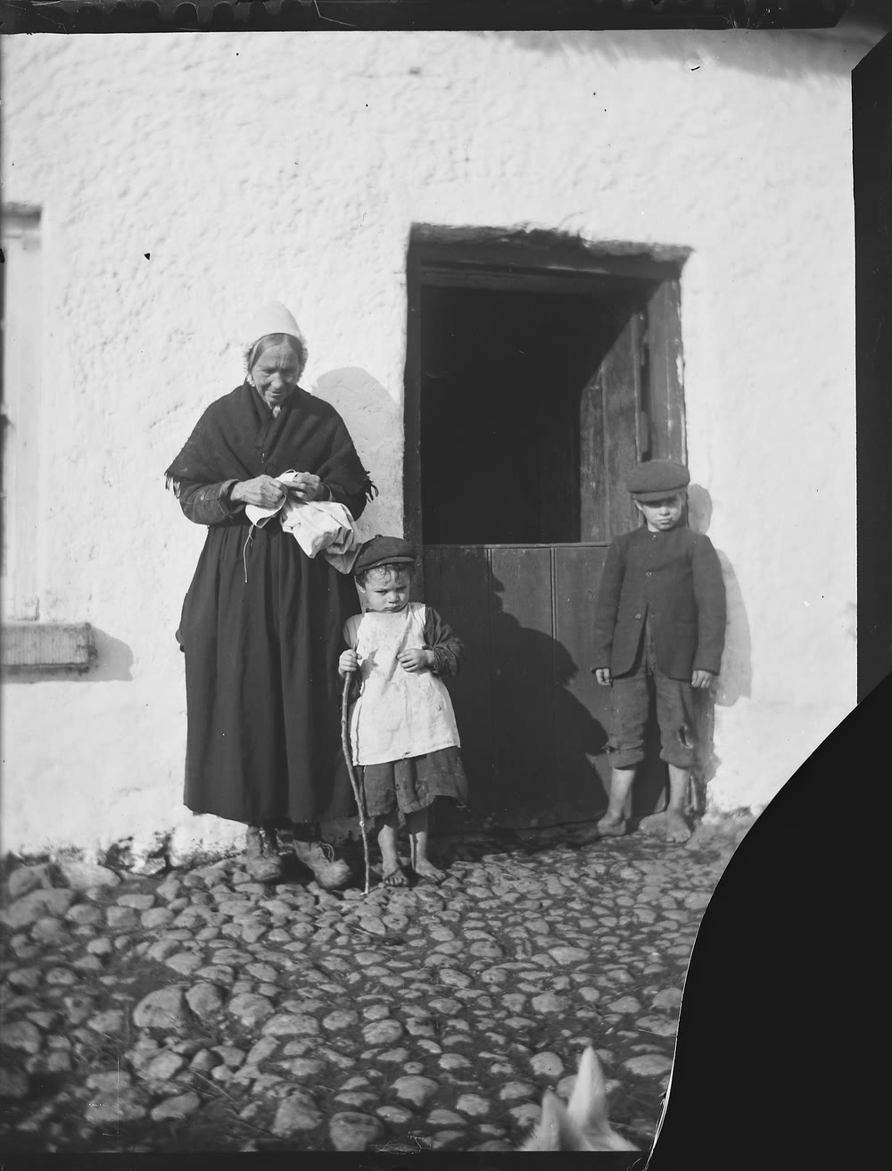 Old photograph of an older woman in a white cap and dark shawl, holding a piece of white cloth, beside the half-door of a whitewashed cottage, with a small barefoot child in a pinafore holding a stick and a barefoot boy in a cap and jacket on the cobbles