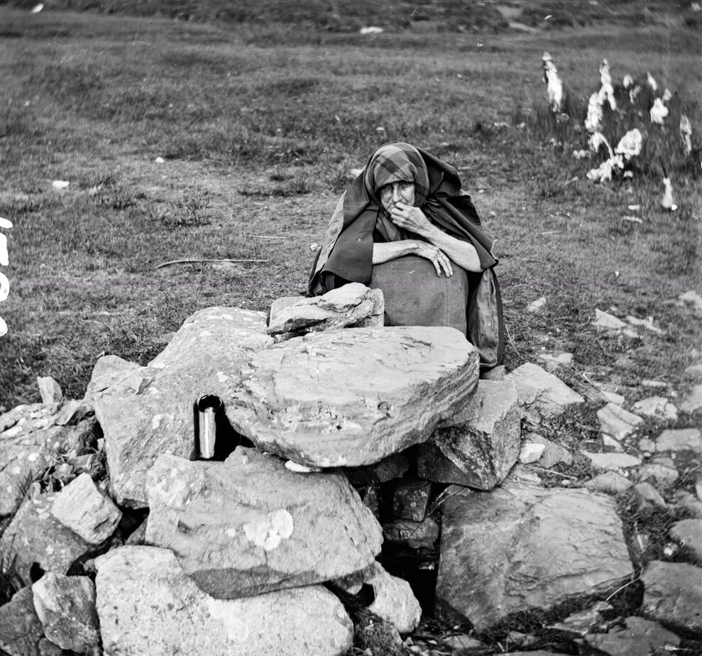 Black-and-white photograph of an elderly woman wrapped in a checked shawl, sitting behind the rough stones of a holy well in a grassy field, with one hand raised to her mouth