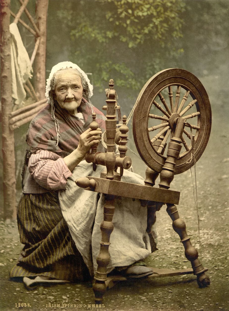 Colour-tinted photograph of an elderly woman in a white frilled cap and striped shawl, seated at a wooden spinning wheel and looking at the camera