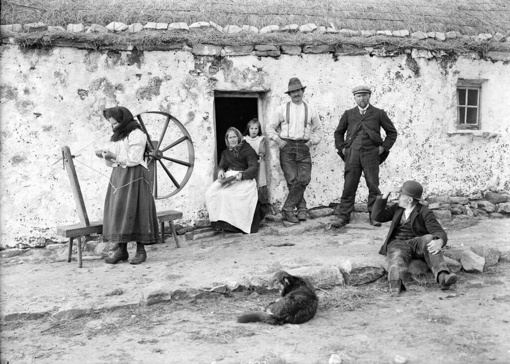 Old photograph of a family outside a whitewashed thatched cottage around 1900: a young woman spins wool at a large walking wheel, an older woman sits in the doorway with wool in her lap beside a girl, two men stand against the wall, another man sits on the stones and a dog lies in the yard