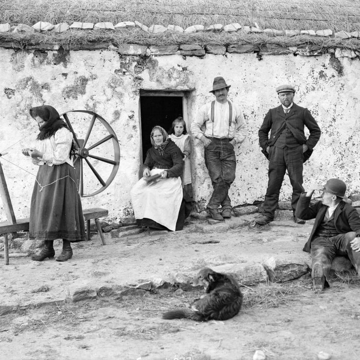 Old photograph of a family outside a whitewashed thatched cottage around 1900: a young woman spins wool at a large walking wheel, an older woman sits in the doorway with wool in her lap beside a girl, two men stand against the wall, another man sits on the stones and a dog lies in the yard