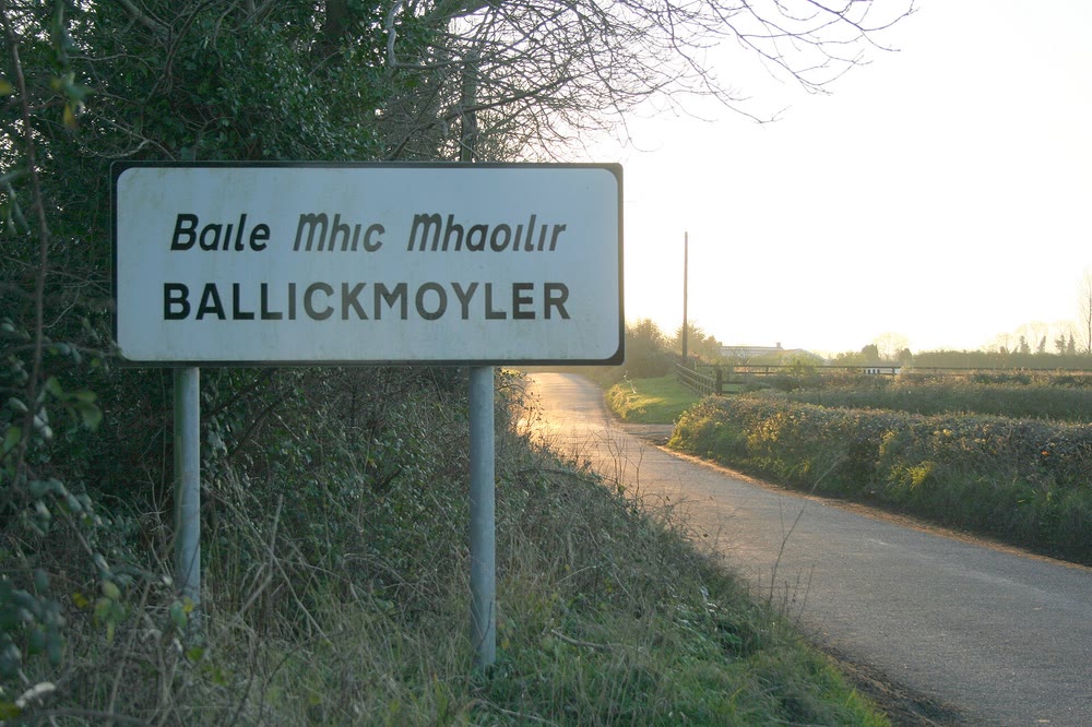 A white roadside sign reading Baile Mhic Mhaoilir in Irish above BALLICKMOYLER in capitals, beside a quiet country road lined with hedges in low winter sunlight