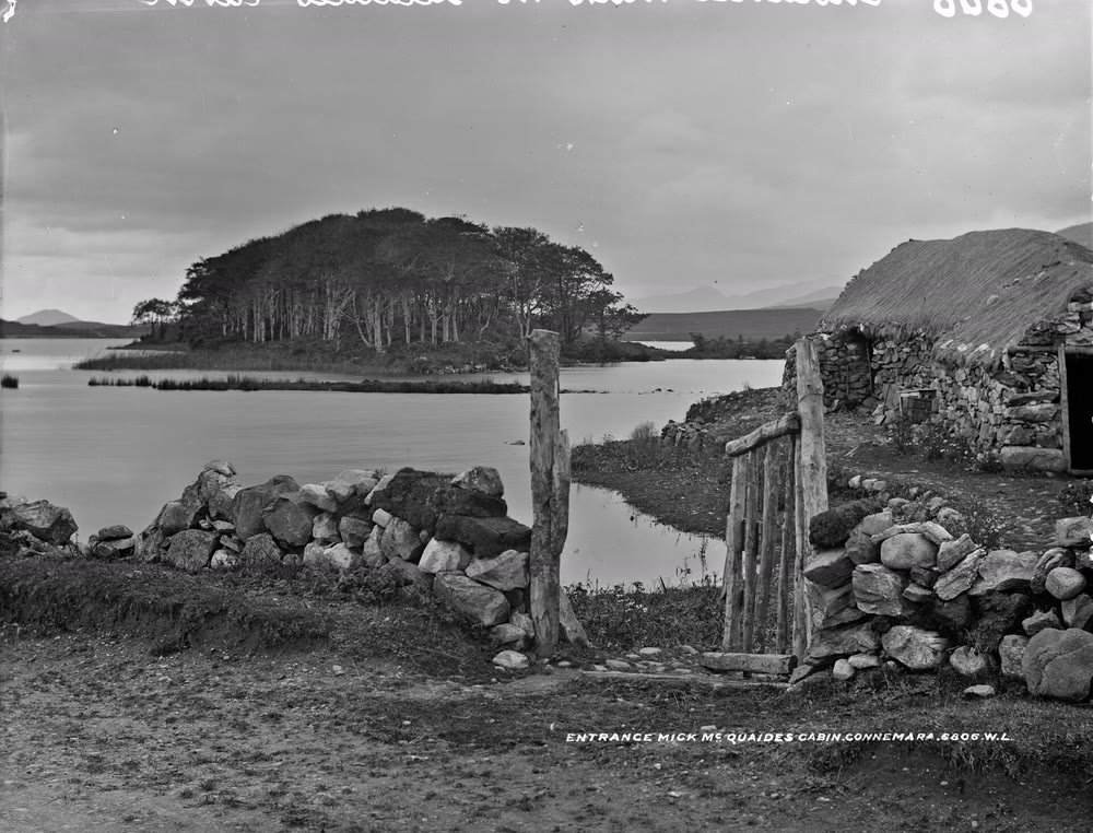Black and white photograph of a thatched stone cabin with a wooden gate beside a lake and a wooded island in Connemara