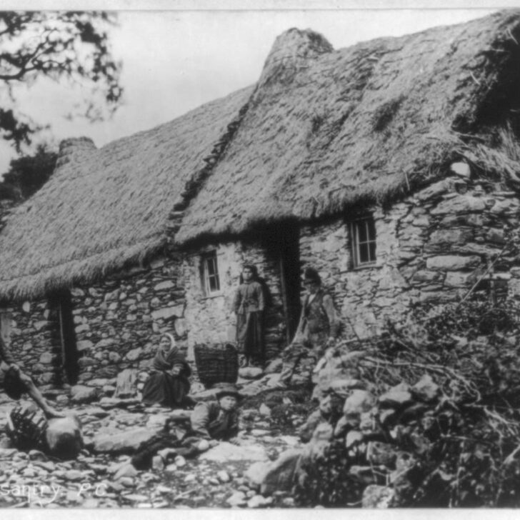 Old photograph of a family gathered outside a long thatched stone cottage on a rocky hillside in Ireland