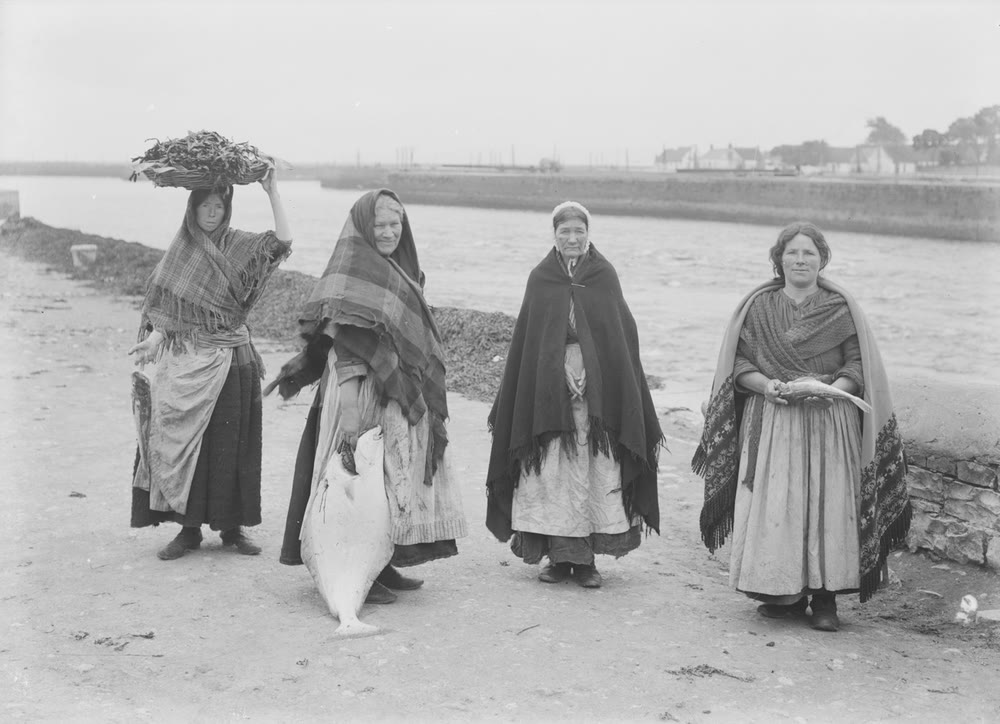 Four women in long shawls and aprons stand on a riverside path in Galway, one balancing a laden basket on her head and another holding a large flat fish, with a quay and low houses across the river behind them
