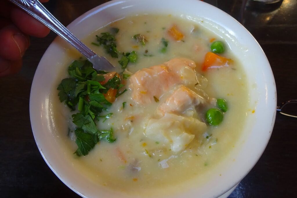 A white bowl of creamy seafood chowder with chunks of salmon and white fish, carrots, peas and fresh parsley, with a spoon resting in it