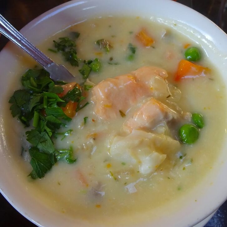 A white bowl of creamy seafood chowder with chunks of salmon and white fish, carrots, peas and fresh parsley, with a spoon resting in it