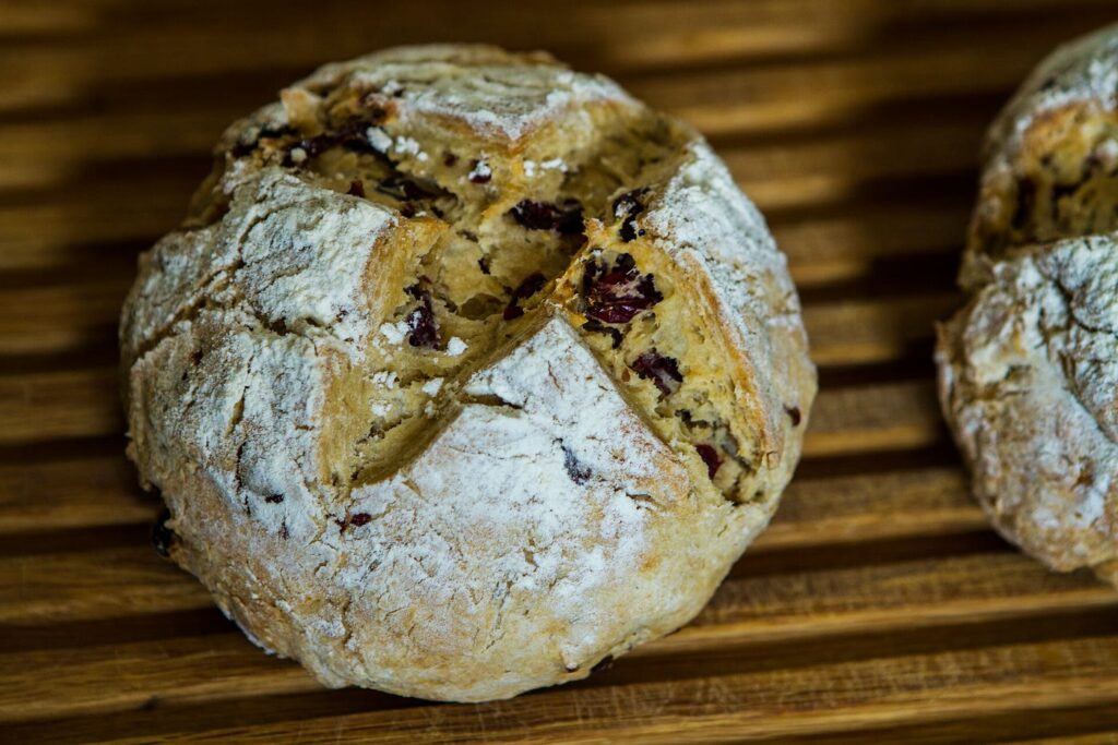A round, flour-dusted Irish soda bread with a deep cross cut into the top, opening to show a soft crumb studded with dried fruit, on a wooden slatted board
