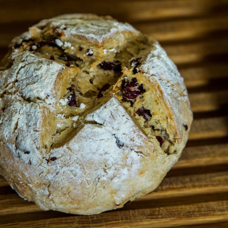 A round, flour-dusted Irish soda bread with a deep cross cut into the top, opening to show a soft crumb studded with dried fruit, on a wooden slatted board