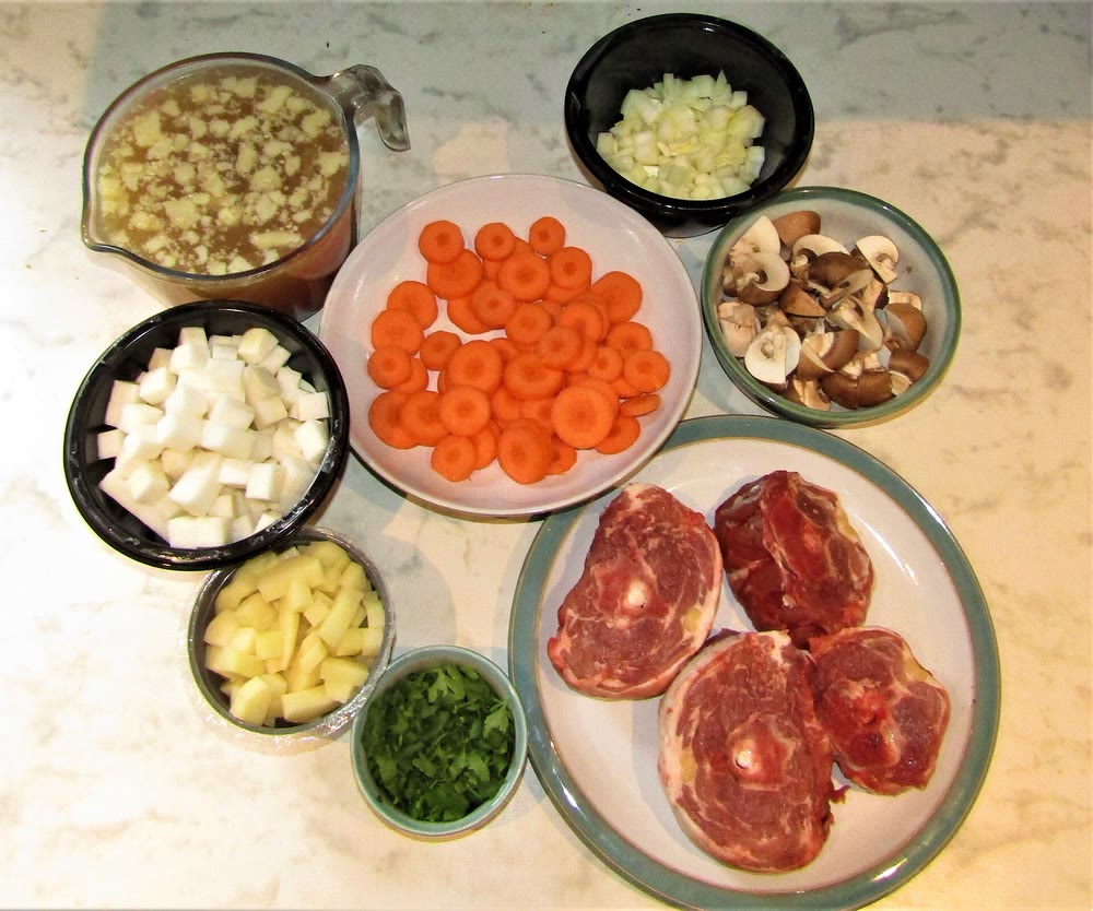Ingredients for Irish stew laid out on a kitchen counter: four round slices of lamb neck on a plate, sliced carrots, diced root vegetables, chopped onion, mushrooms, parsley and a jug of broth