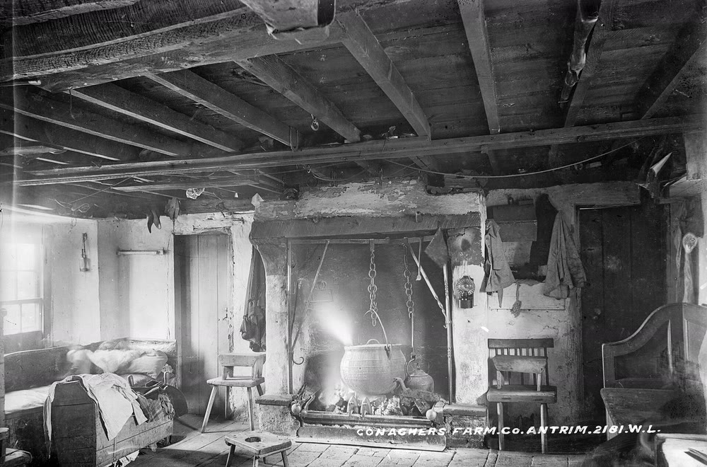 An Irish farmhouse kitchen around the 1890s: a black pot hangs on chains over an open fire in a wide stone hearth, with a kettle, stools, a chair and a settle around it and rafters overhead