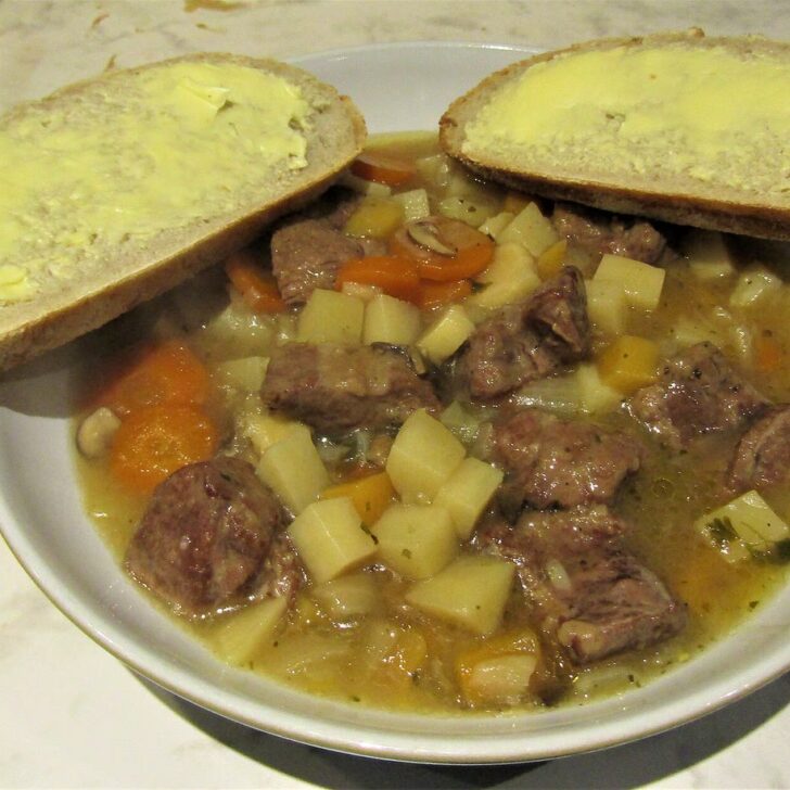 A white bowl of Irish stew with chunks of lamb, diced potato and carrot in a golden broth, with two slices of buttered bread resting on the rim
