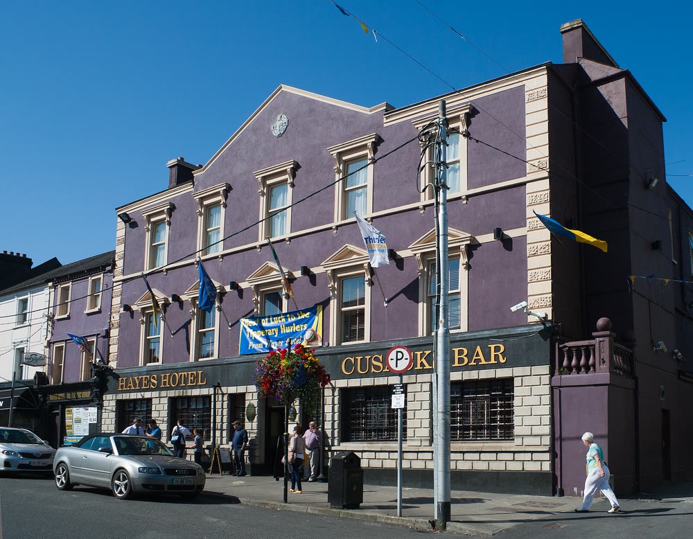 Three-storey purple-painted hotel on a sunny street corner, with Hayes Hotel and Cusack Bar written in gold above the ground floor, flags and a hurling banner on the front and a few people on the footpath