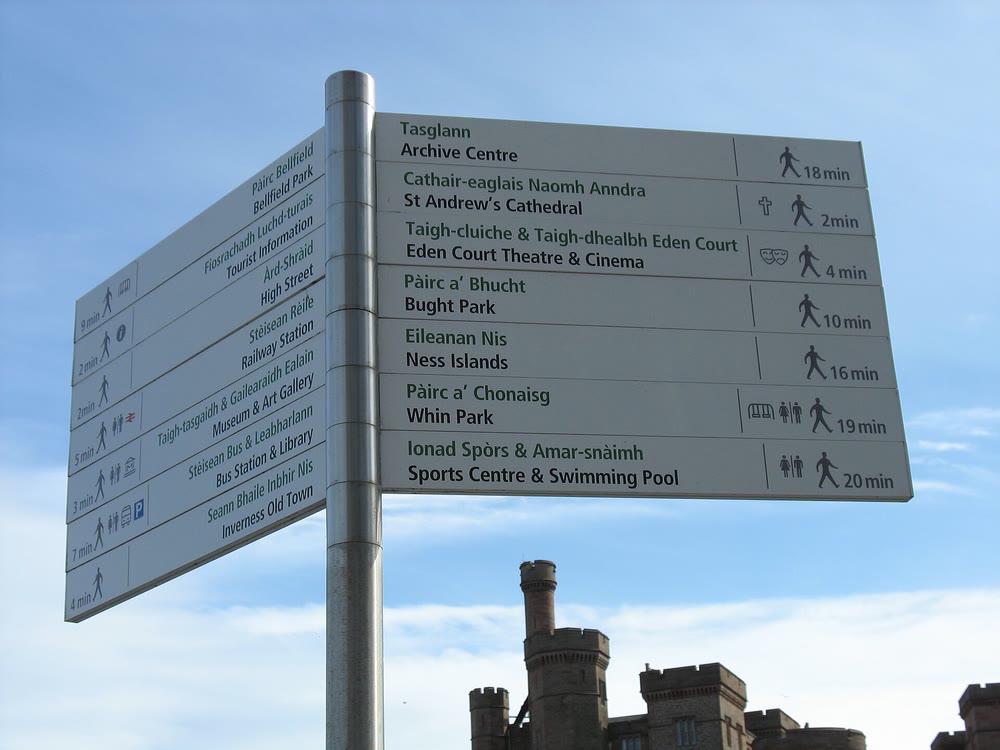 Metal signpost against a blue sky with white finger signs in Scottish Gaelic and English, including &Agrave;rd-Shr&agrave;id for High Street, St&egrave;isean R&egrave;ile for Railway Station and P&agrave;irc a' Bhucht for Bught Park, with a castle turret below
