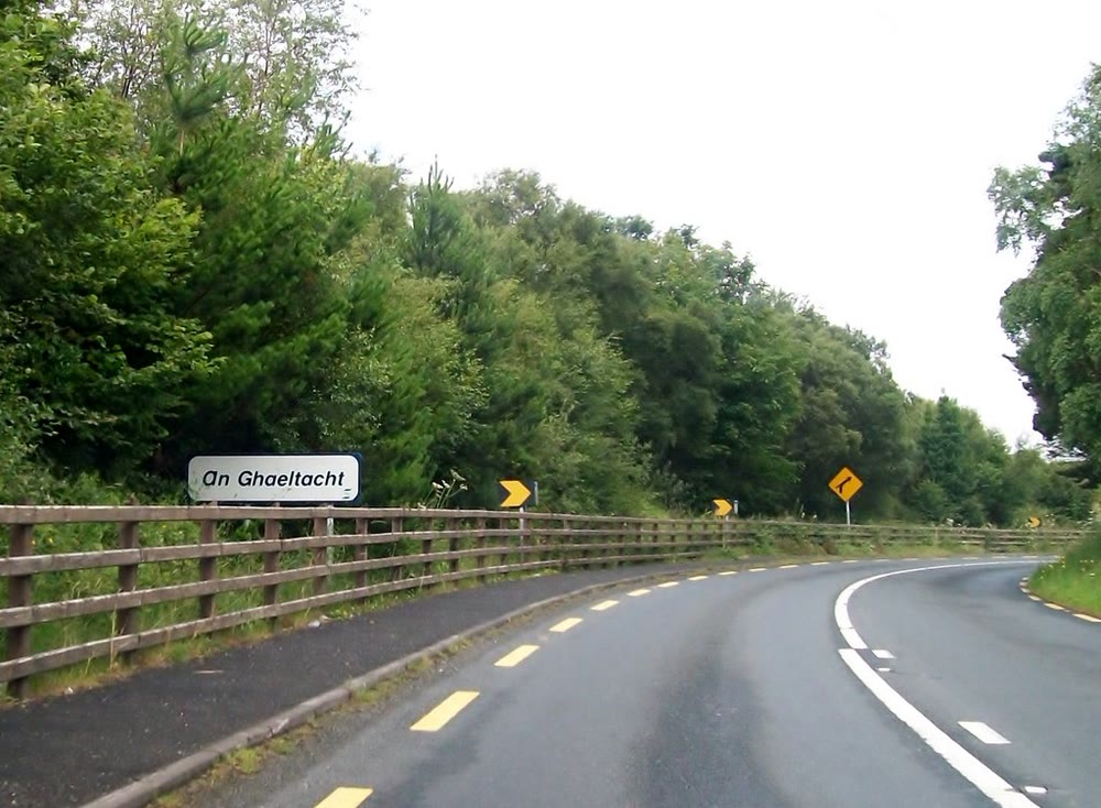 White roadside sign reading An Ghaeltacht beside a wooden fence on a curving road through thick green trees, with yellow bend warning signs ahead