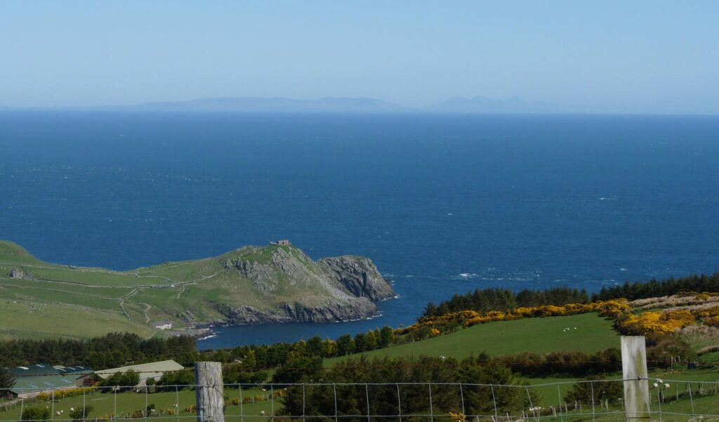 Green headland of Torr Head with a small ruined building on its summit, a deep blue sea beyond and the faint hills of Scotland along the horizon, with fields, gorse in yellow flower and a wire fence in the foreground