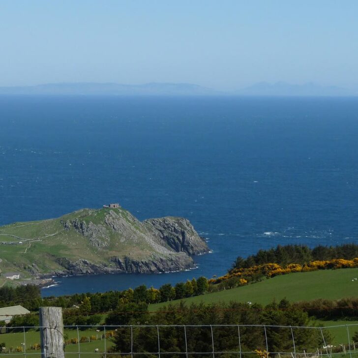 Green headland of Torr Head with a small ruined building on its summit, a deep blue sea beyond and the faint hills of Scotland along the horizon, with fields, gorse in yellow flower and a wire fence in the foreground