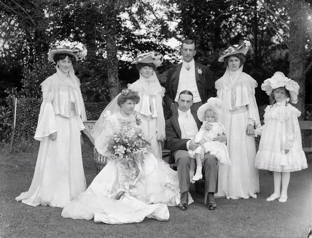 1903 black-and-white wedding group in a garden: the bride sits with a large bouquet and a long train beside a seated man holding a small child, with bridesmaids in wide flowered hats standing behind