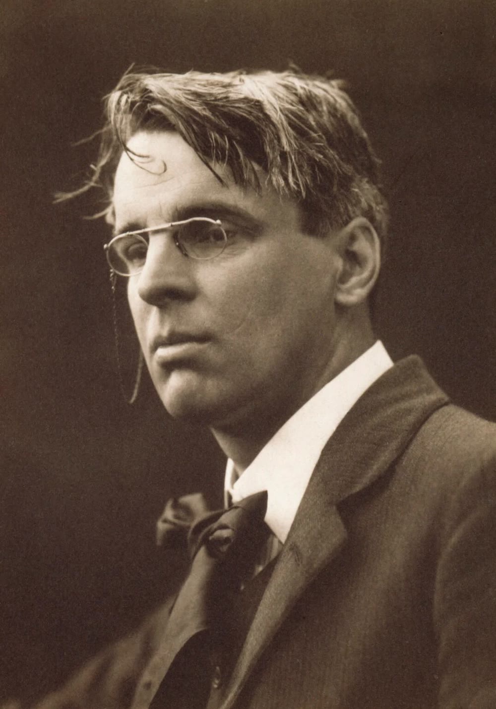 Sepia portrait of the poet W. B. Yeats in 1911, with untidy swept-back hair and pince-nez glasses on a cord, wearing a dark jacket and loose tie