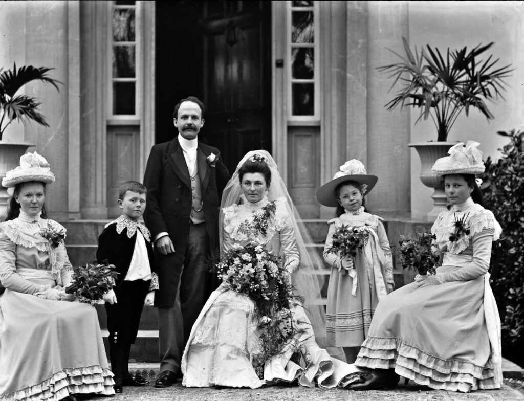 1900 wedding photograph on the steps of a house in Clonmel: a bride in a long veil holding a large bouquet sits beside the standing groom, flanked by bridesmaids in wide hats, a young bridesmaid and a small page boy