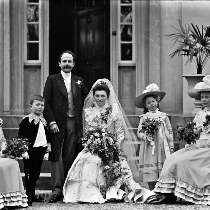 1900 wedding photograph on the steps of a house in Clonmel: a bride in a long veil holding a large bouquet sits beside the standing groom, flanked by bridesmaids in wide hats, a young bridesmaid and a small page boy