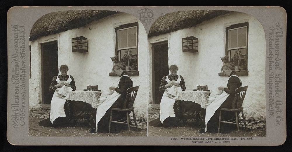 A 1904 stereograph of two women in white aprons sewing lace at a table outside a whitewashed thatched cottage