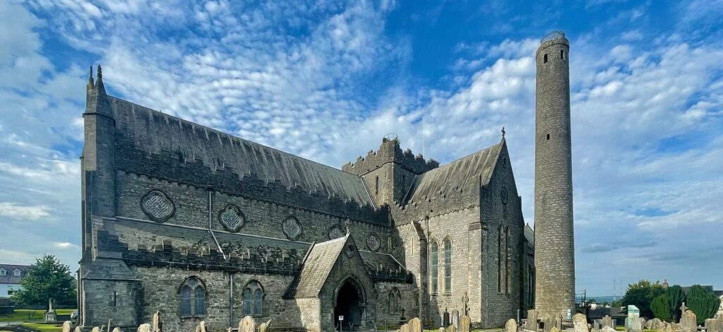 St Canice's Cathedral in Kilkenny: a long grey limestone medieval church with stepped battlements and a squat central tower, a tall round tower beside it, and old headstones on the lawn in front under a blue sky