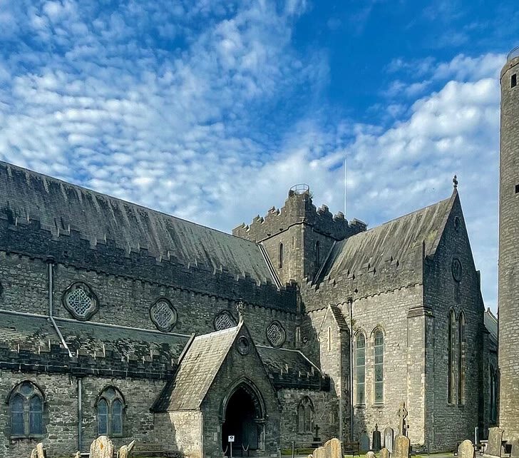 St Canice's Cathedral in Kilkenny: a long grey limestone medieval church with stepped battlements and a squat central tower, a tall round tower beside it, and old headstones on the lawn in front under a blue sky
