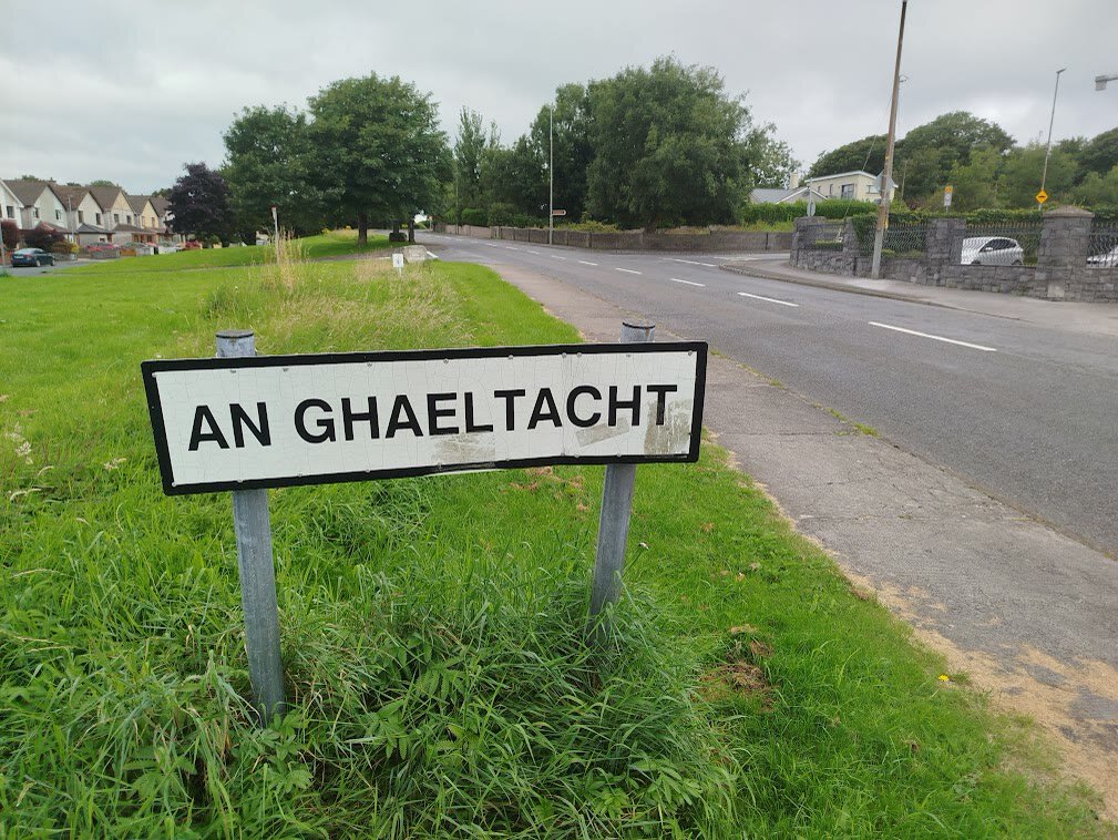 White road sign reading An Ghaeltacht on a grassy verge beside a suburban road in Mionlach (Menlo), Galway, with houses and trees behind