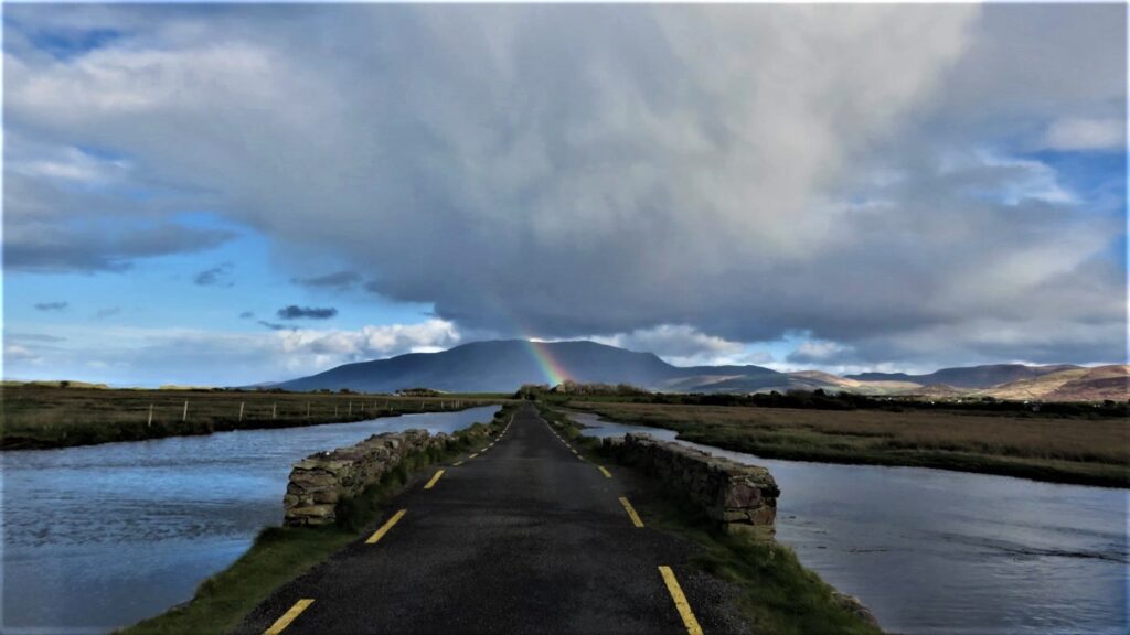 A rainbow over the Slieve Mish mountains under a dark shower cloud, seen down a narrow causeway road between tidal water near Castlegregory, Co. Kerry