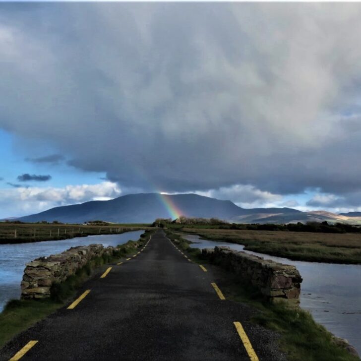 A rainbow over the Slieve Mish mountains under a dark shower cloud, seen down a narrow causeway road between tidal water near Castlegregory, Co. Kerry