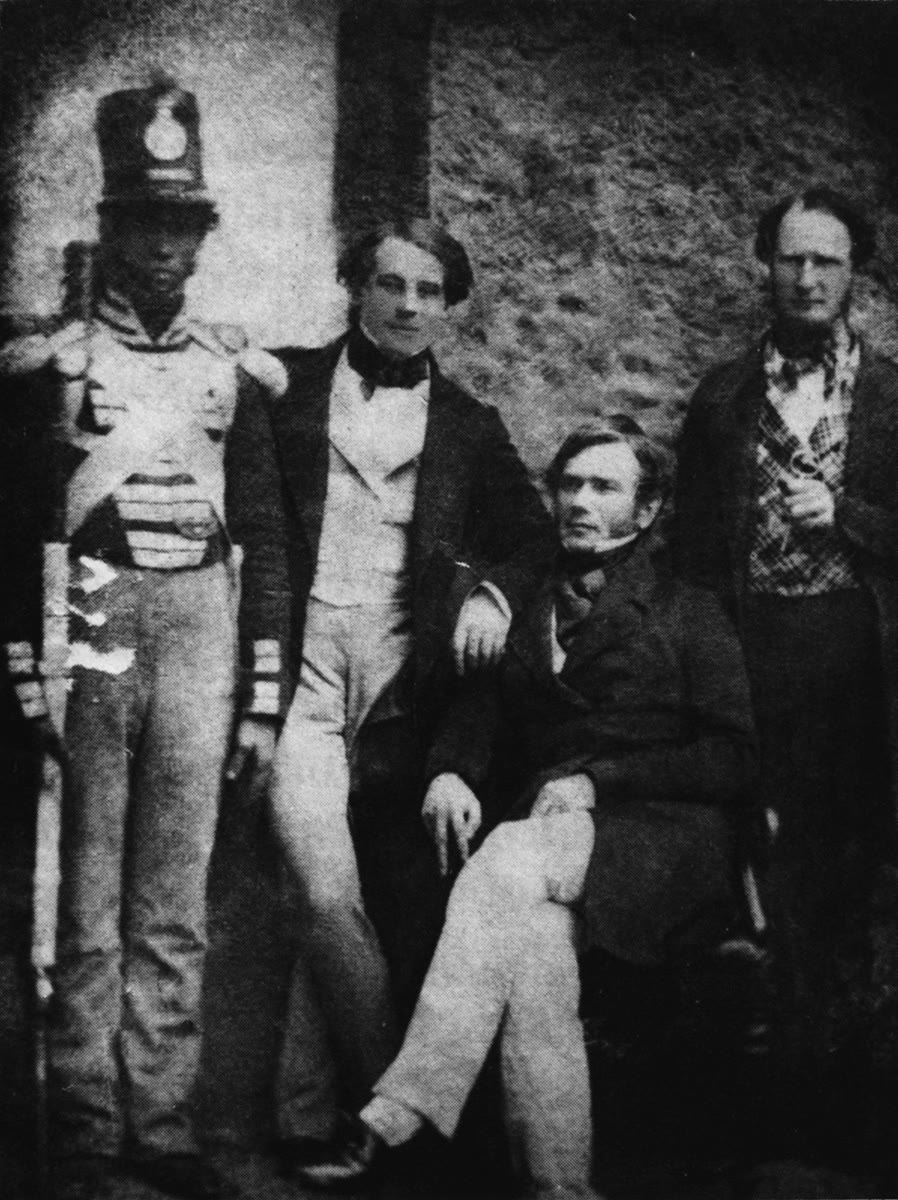 1848 photograph of four men against a stone wall: a soldier in a tall shako, a young man in a dark coat and pale waistcoat, a seated man with his legs crossed, and a man in a checked waistcoat holding a pipe