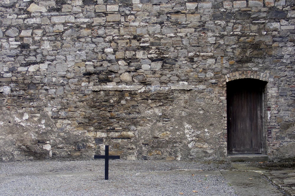 A plain black cross on the gravel of the Stonebreakers' Yard in front of a high rubble-stone wall, with an arched wooden door on the right