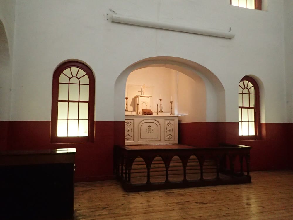 Prison chapel with dark red lower walls, white upper walls and two arched windows, and a pale altar marked IHS in an arched alcove behind a low wooden rail