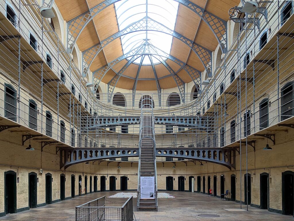 The Victorian East Wing of Kilmainham Gaol: three tiers of cell doors and iron walkways around an open hall, with a central iron staircase rising towards a curved glass and timber skylight