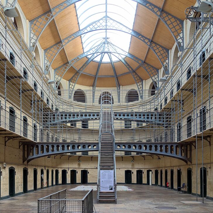 The Victorian East Wing of Kilmainham Gaol: three tiers of cell doors and iron walkways around an open hall, with a central iron staircase rising towards a curved glass and timber skylight