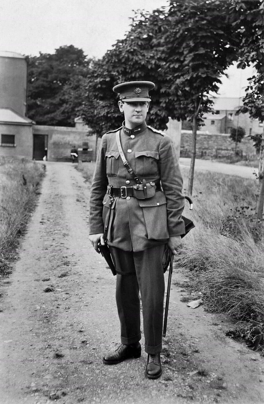 Michael Collins in 1922, standing on a dirt path in a peaked cap and army uniform with a Sam Browne belt, holding a cane, with young trees and a stone wall behind him