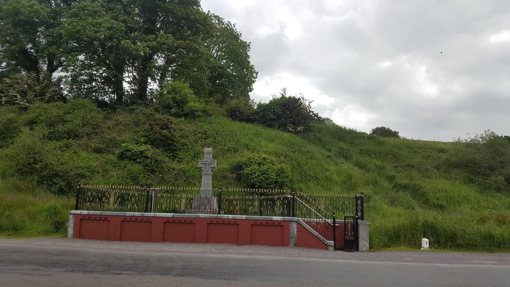 A stone Celtic cross inside a railed enclosure on a red-painted wall at the roadside at Béal na Bláth, with a steep green hillside and trees behind