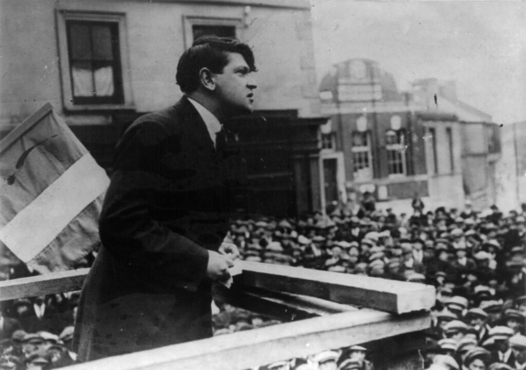 Michael Collins in a dark suit leans on a wooden platform rail and speaks to a packed crowd of men in caps, with a tricolour flag at the left and town buildings behind