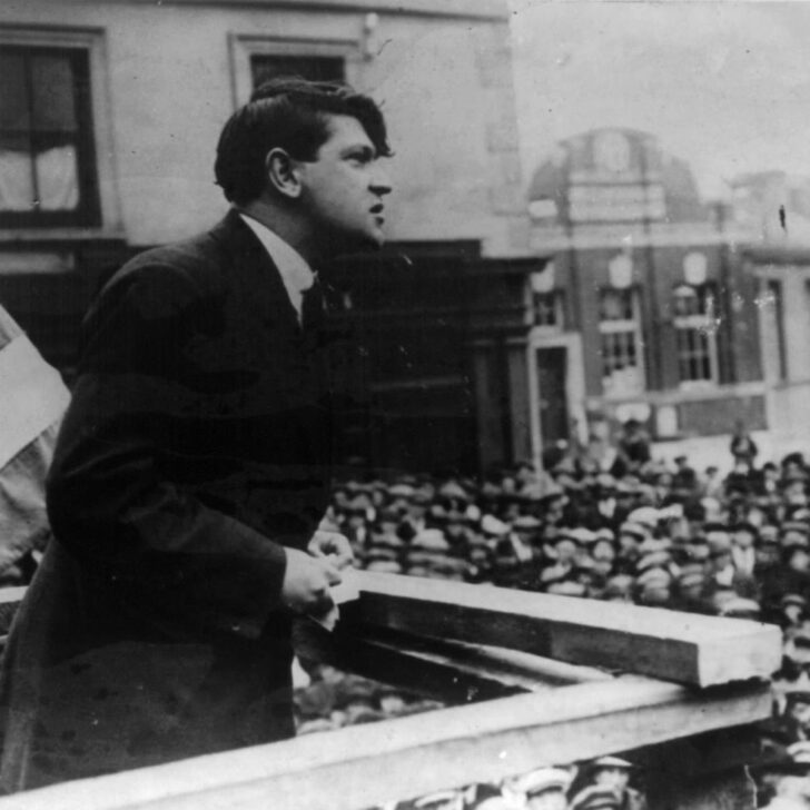 Michael Collins in a dark suit leans on a wooden platform rail and speaks to a packed crowd of men in caps, with a tricolour flag at the left and town buildings behind