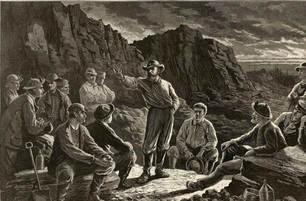 Molly Maguires meeting: 1874 Harper's Weekly engraving of Pennsylvania coal miners gathered among rocks at dusk, listening to a bearded man in a wide-brimmed hat