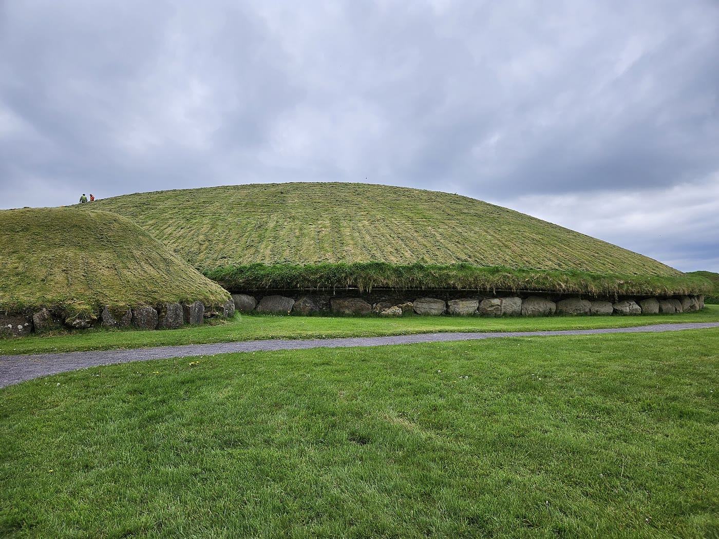 Knowth's huge grassy passage tomb ringed by kerbstones, with a smaller satellite mound beside it under a grey sky