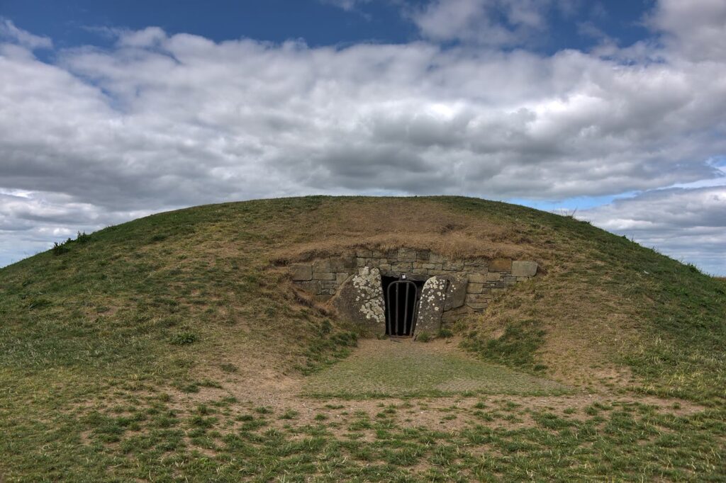 The Mound of the Hostages on the Hill of Tara: a grassy Neolithic passage tomb mound with a small barred stone entrance under a big cloudy sky